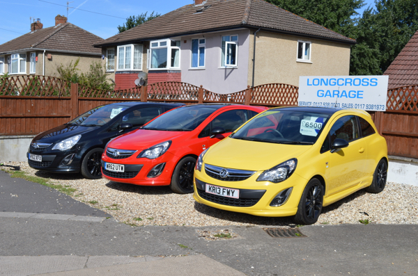 Three Vauxhall hatchbacks, black, red and yellow, in front of the Longcross sign