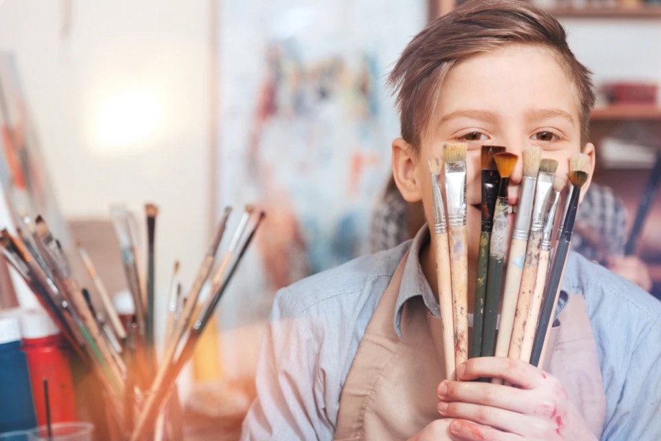A boy in an art apron holding a handful of paintbrushes up to his face