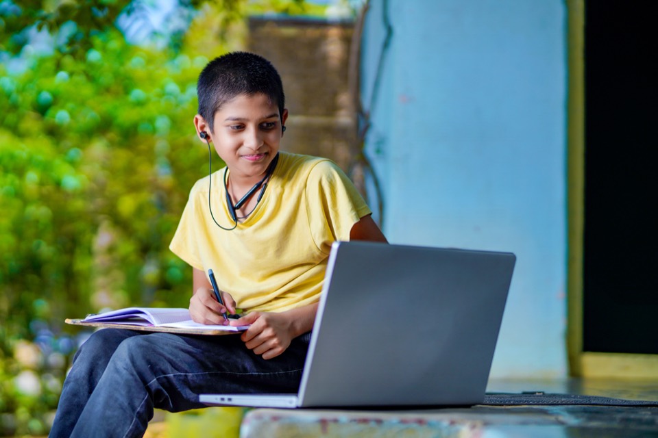 A boy in a yellow T-shirt writing in his notebook beside his laptop