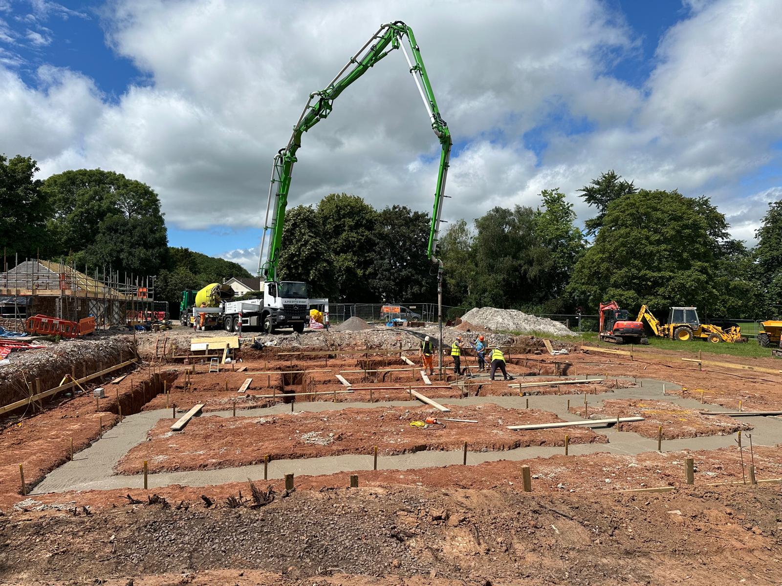 A concrete pump's boom placing concrete into strip footings, a green and yellow mixer behind it
