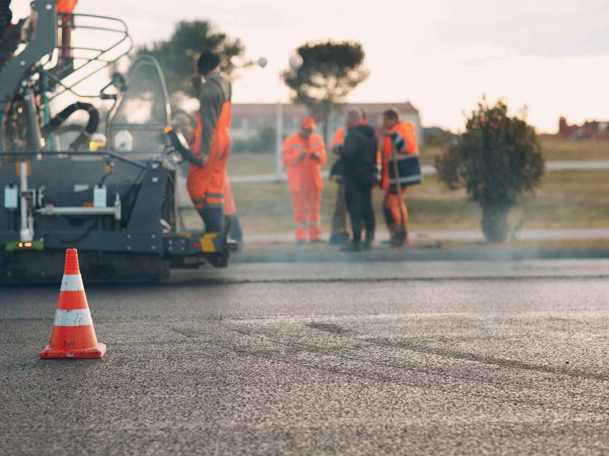 A road crew and paver laying a new asphalt surface behind traffic cones
